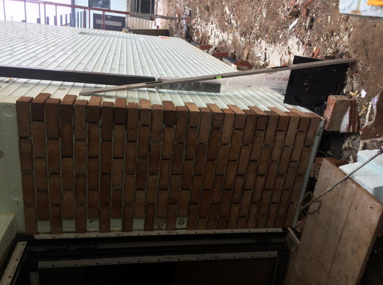 Fragment ściany budynku w trakcie prac elewacyjnych: widoczna warstwa izolacji termicznej z białego styropianu oraz częściowo ułożona okładzina z cegły klinkierowej w kolorze brązowym, z poziomnicą...