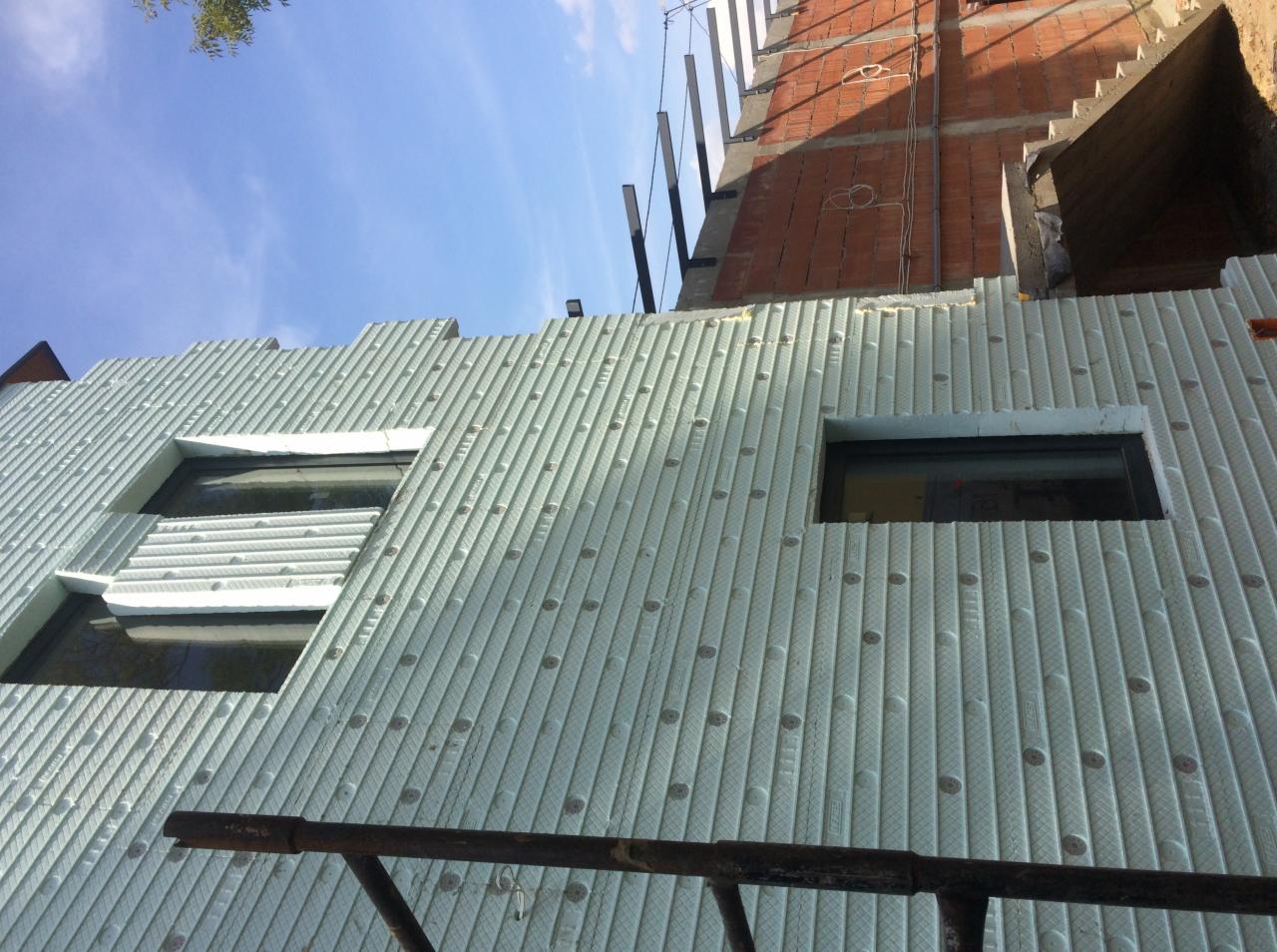 Ściana budynku w trakcie prac izolacyjnych, widoczne płyty styropianowe z charakterystycznym wzorem i otwory montażowe, okna częściowo zakryte izolacją, w tle surowa ceglana ściana w trakcie budowy.