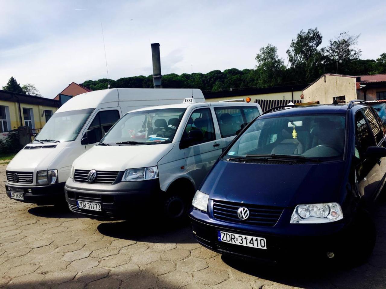 Trzy zaparkowane pojazdy: biały bus dostawczy, biały van z napisem 'TAXI' na dachu i granatowy samochód osobowy typu minivan, wszystkie z polskimi tablicami rejestracyjnymi, stojące na brukowanym...