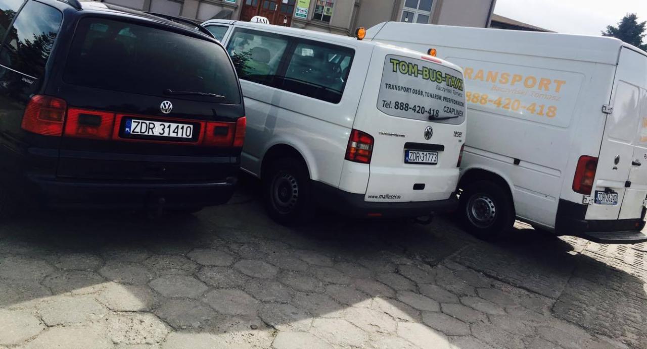 Trzy busy transportowe Volkswagen zaparkowane obok siebie na brukowanej ulicy, z widocznymi numerami rejestracyjnymi i oznaczeniami firmowymi na dwóch z nich, w tym 'TOM-BUS-TAXI' z numerem telefonu.