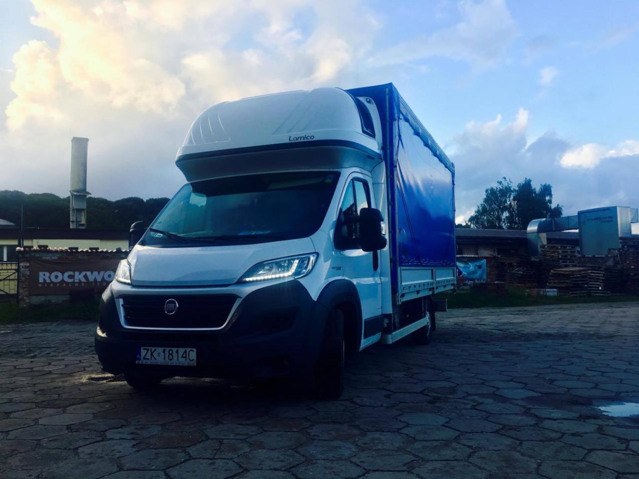 Biały samochód dostawczy z niebieską plandeką na parkingu, widoczny numer rejestracyjny ZK 1814C, w tle budynki i zachmurzone niebo.
