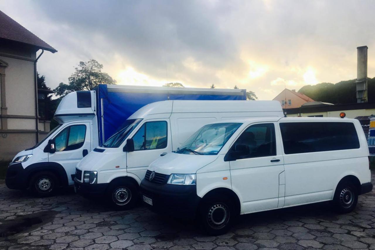 Trzy białe samochody dostawcze różnych typów zaparkowane obok siebie na brukowanym placu, widoczne budynki w tle, pochmurne niebo.