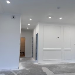 WIERZBIAK Marek Wierzbiak - Wnętrze w trakcie prac wykończeniowych, świeżo pomalowane na biało ściany z dekoracyjnymi panelami, widoczne punkty oświetleniowe w suficie i surowa betonowa podłoga zabezpieczona folią malarską.