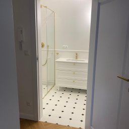 WIERZBIAK Marek Wierzbiak - Elegancka łazienka w Zabrzu po remoncie: białe płytki z czarnymi akcentami, kabina prysznicowa ze złotymi detalami, biała szafka pod umywalką z trzema szufladami.