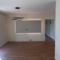 WIERZBIAK Marek Wierzbiak - Jasne wnętrze po wykończeniu: panele podłogowe, ściana z podświetleniem LED i dekoracyjnymi lamelami, nowoczesna lampa wisząca. Widok na sąsiednie pomieszczenie.