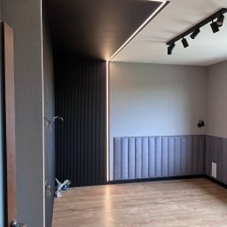 WIERZBIAK Marek Wierzbiak - Nowoczesne wnętrze z czarną ścianą z lameli i podświetleniem LED, szara tapeta, panele podłogowe i oświetlenie punktowe na suficie. Częściowo widoczne kable.