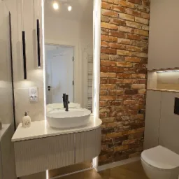 Nowoczesna łazienka z umywalką nablatową, lustrem z podświetleniem LED i ścianą z cegły. Parkiet w jodełkę, minimalistyczna armatura i ciepła kolorystyka.