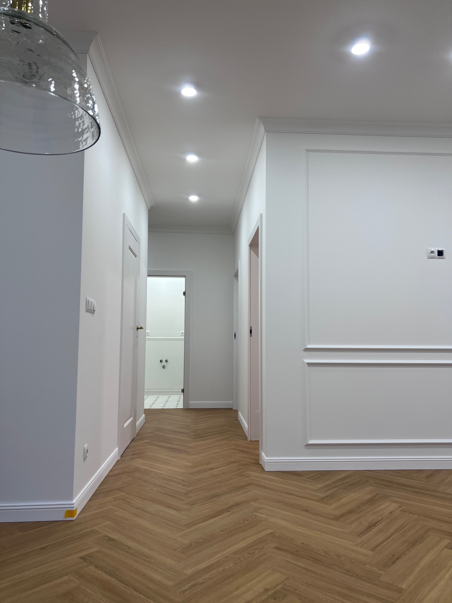 Korytarz po remoncie z parkietem ułożonym w jodełkę, białe ściany z dekoracyjnymi listwami, oświetlenie sufitowe punktowe, widok na otwarte drzwi do łazienki z biało-czarną podłogą.