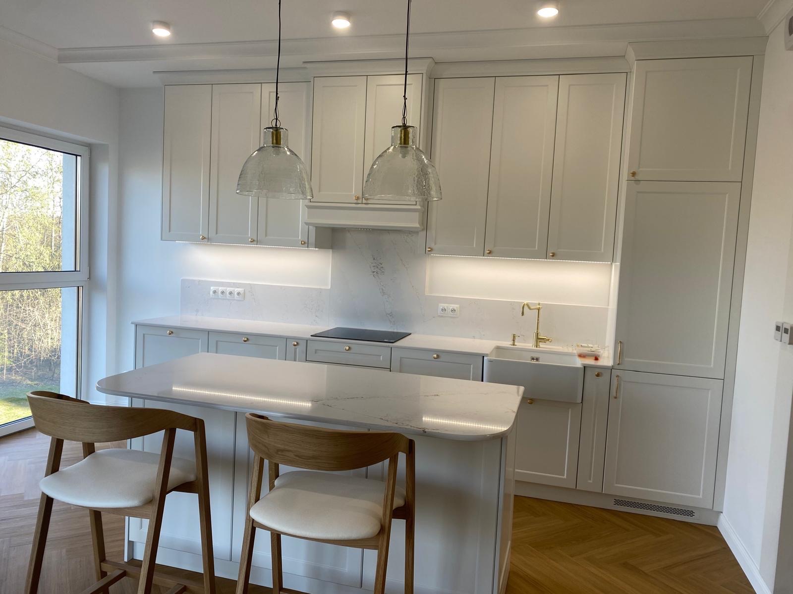 Jasna kuchnia w stylu nowoczesnym: białe szafki z subtelnymi złotymi uchwytami, marmurowy blat, wyspa z hokerami i designerskie lampy wiszące.