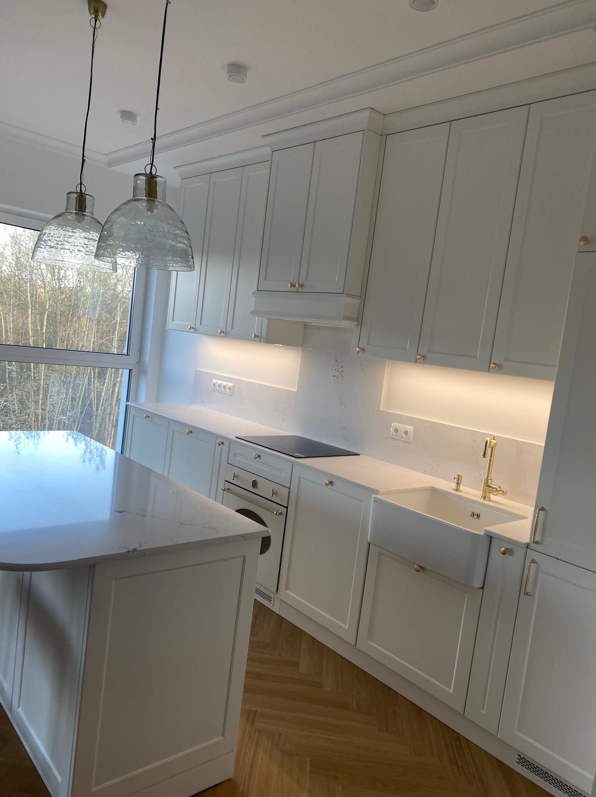 Jasna kuchnia w stylu klasycznym z wyspą, drewnianą podłogą i oświetleniem LED pod szafkami. Widoczne okno z widokiem na drzewa.