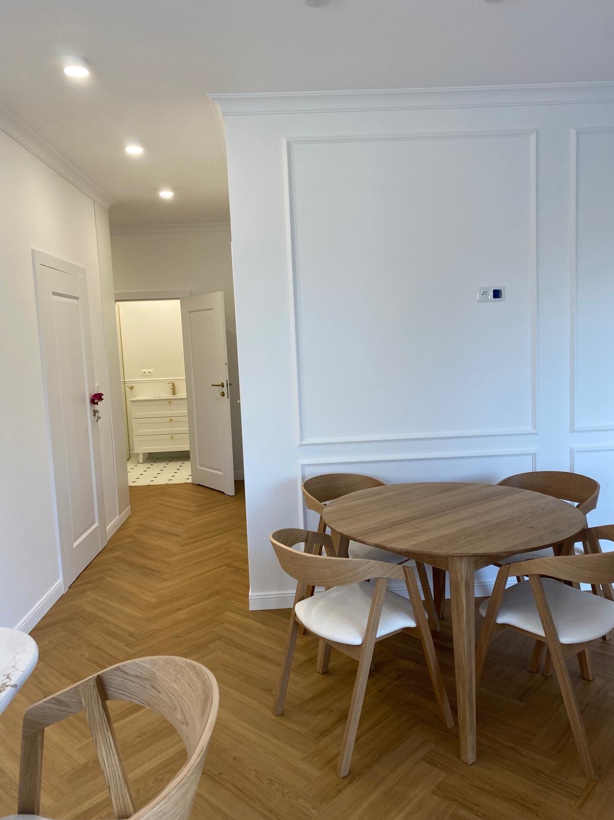 Jasne wnętrze z drewnianą podłogą w jodełkę, okrągłym stołem i krzesłami. W tle widoczna łazienka z białą komodą, dostępna przez otwarte drzwi.