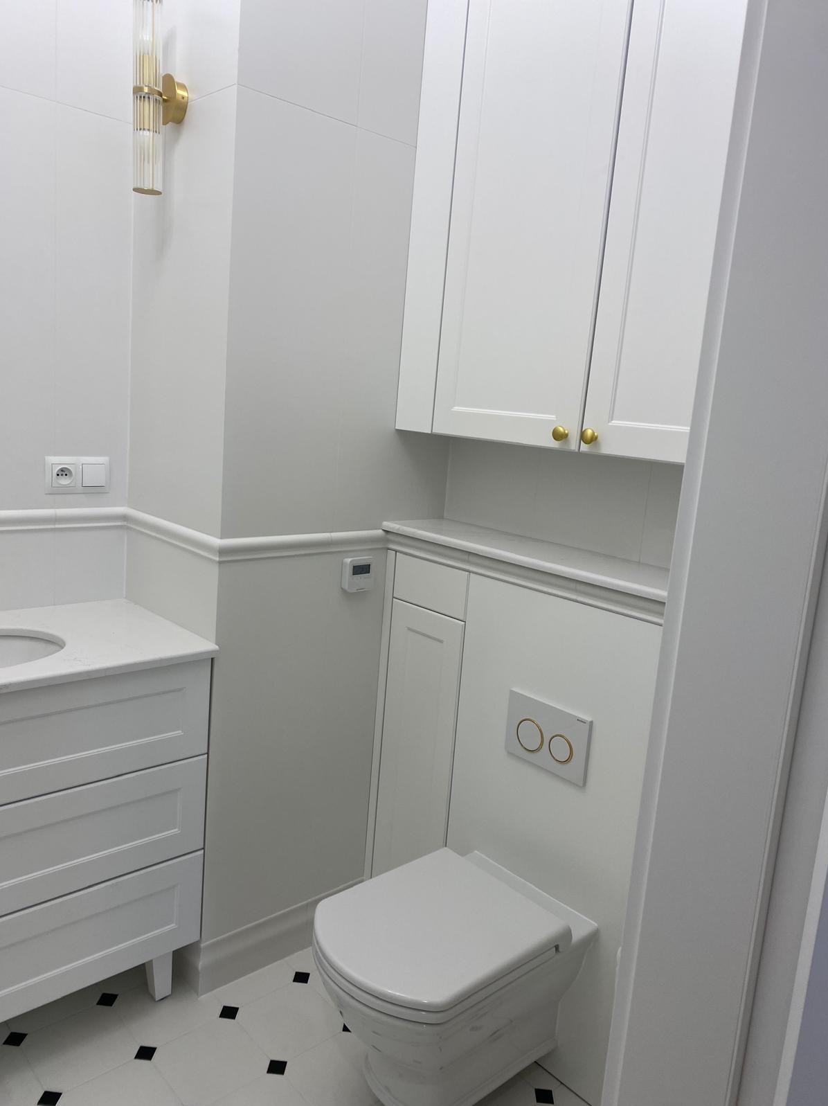Biała łazienka z toaletą, umywalką z szafką i wiszącą szafką. Na ścianie złoty kinkiet i przycisk spłuczki w tym samym kolorze. Czarno-biała podłoga.