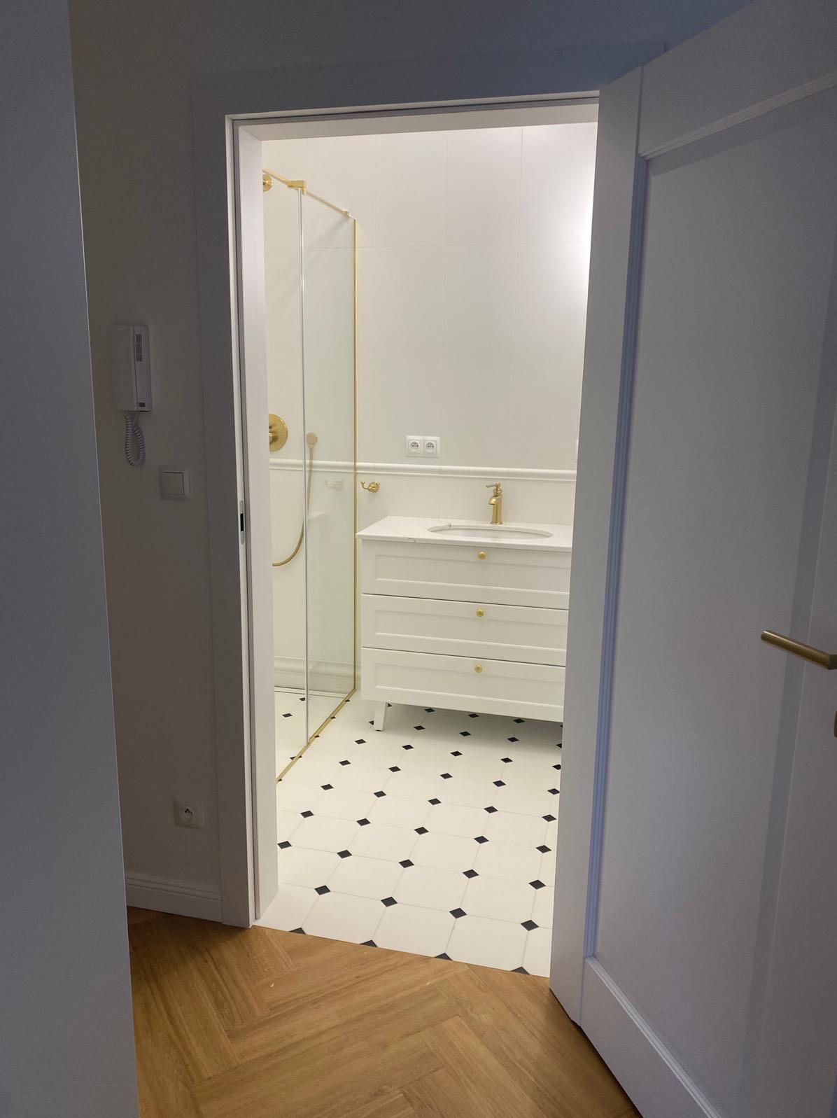 Elegancka łazienka w Zabrzu po remoncie: białe płytki z czarnymi akcentami, kabina prysznicowa ze złotymi detalami, biała szafka pod umywalką z trzema szufladami.