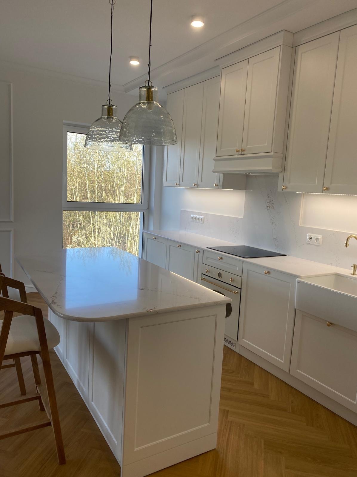Jasna kuchnia z wyspą, białe szafki z złotymi uchwytami, marmurowy blat i parkiet w jodełkę. Nad wyspą wiszą dwie szklane lampy. Za oknem widok na drzewa.