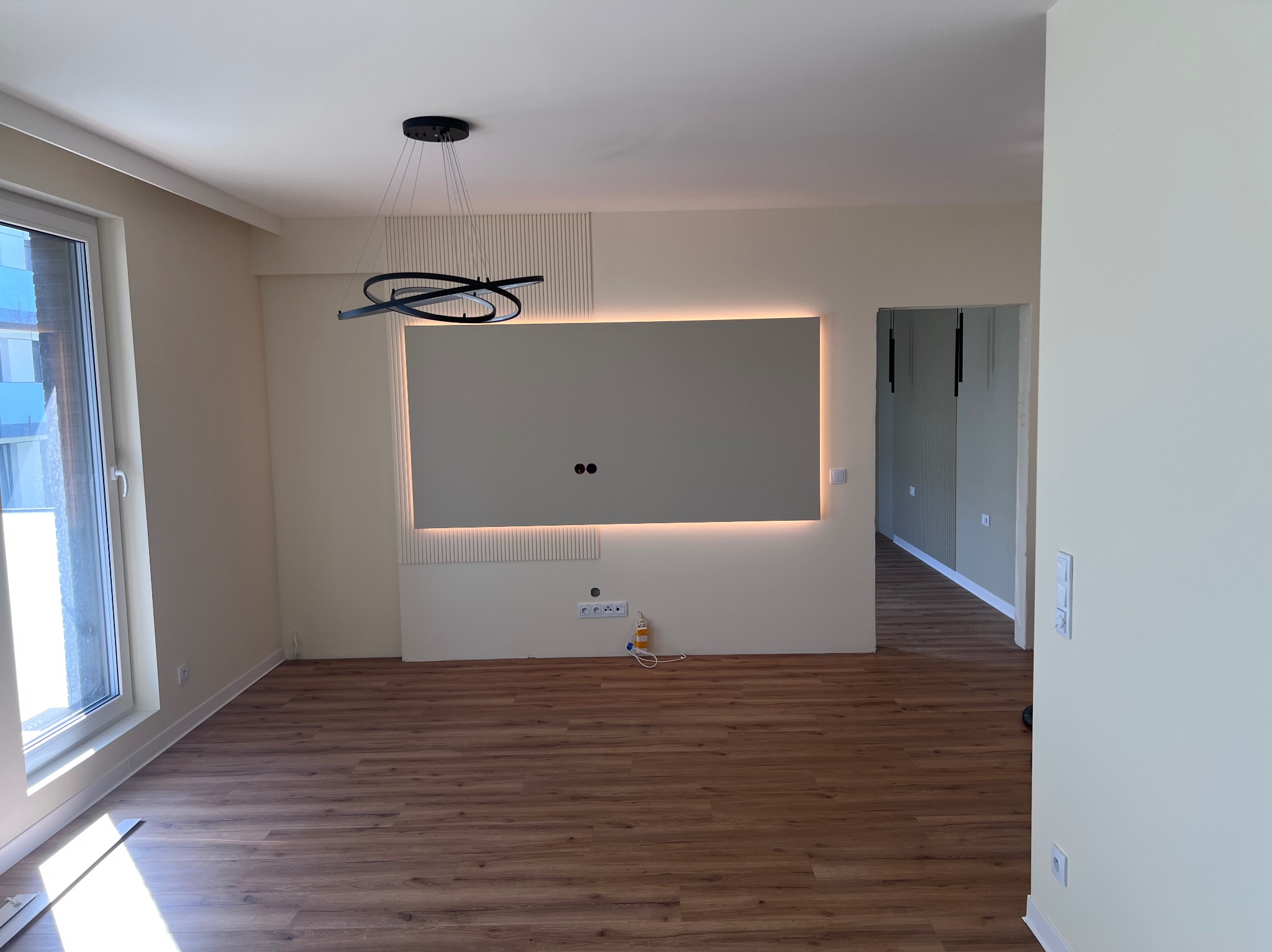 Jasne wnętrze po wykończeniu: panele podłogowe, ściana z podświetleniem LED i dekoracyjnymi lamelami, nowoczesna lampa wisząca. Widok na sąsiednie pomieszczenie.