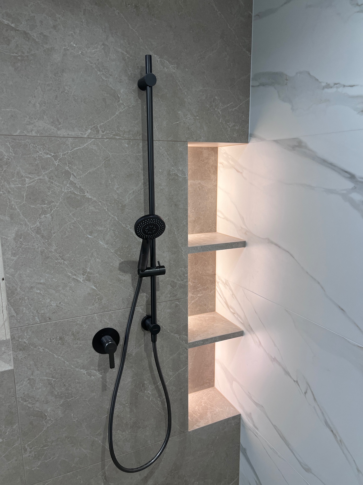 Nowoczesna kabina prysznicowa z czarną armaturą i podświetlanymi półkami wnękowymi, wykończona płytkami imitującymi marmur.