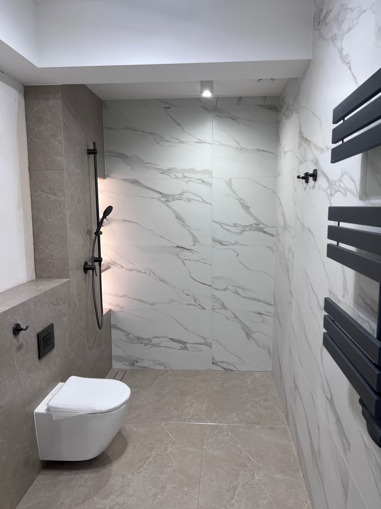 Nowoczesna łazienka z marmurowymi płytkami, wiszącą toaletą i czarną armaturą. Minimalistyczny design, jasne wnętrze z akcentami szarości i bieli.
