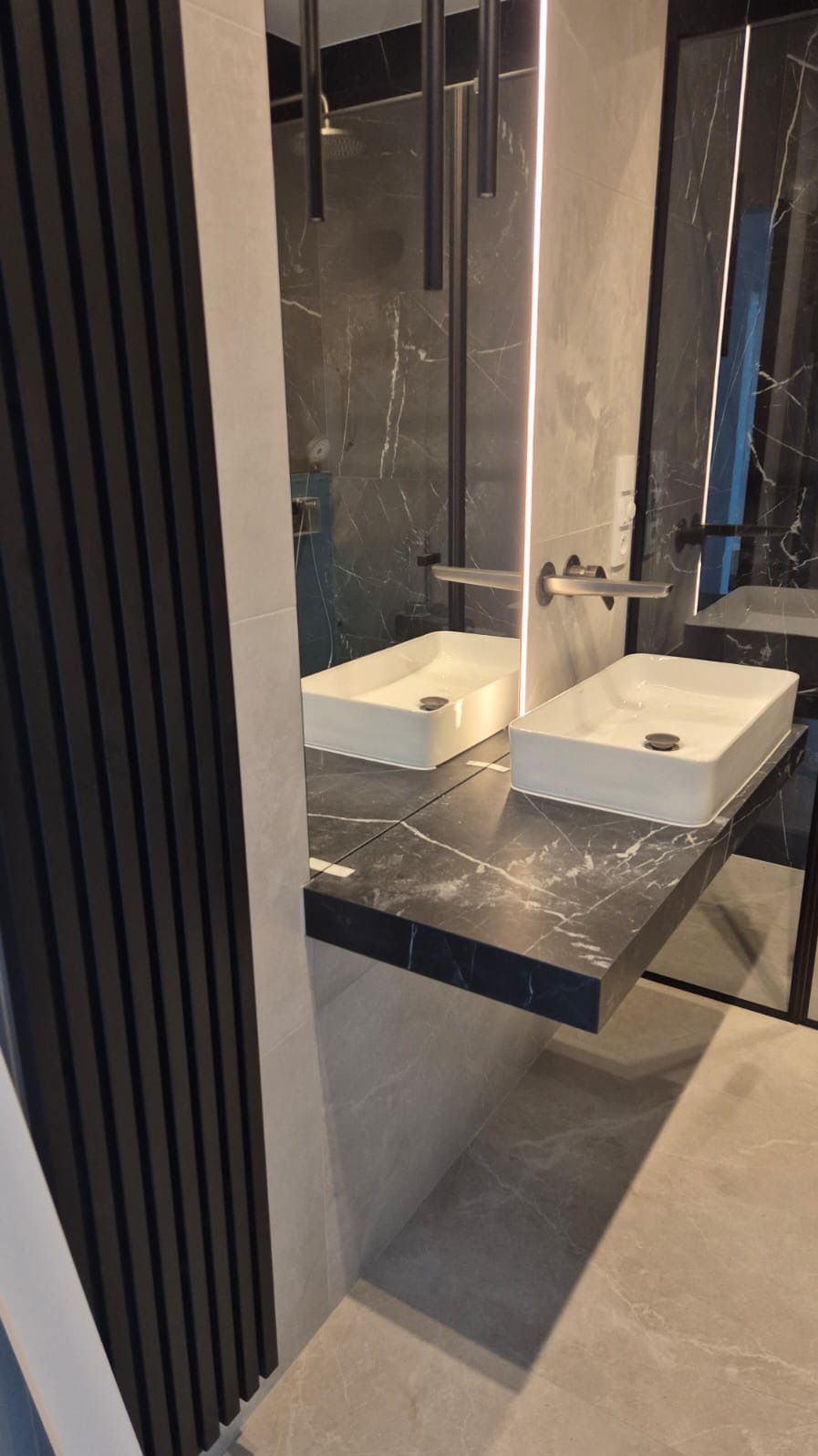 Nowoczesna łazienka z dwoma białymi umywalkami na blacie z czarnego marmuru, oświetlona listwą LED, z elementami dekoracyjnymi w kolorze grafitowym.