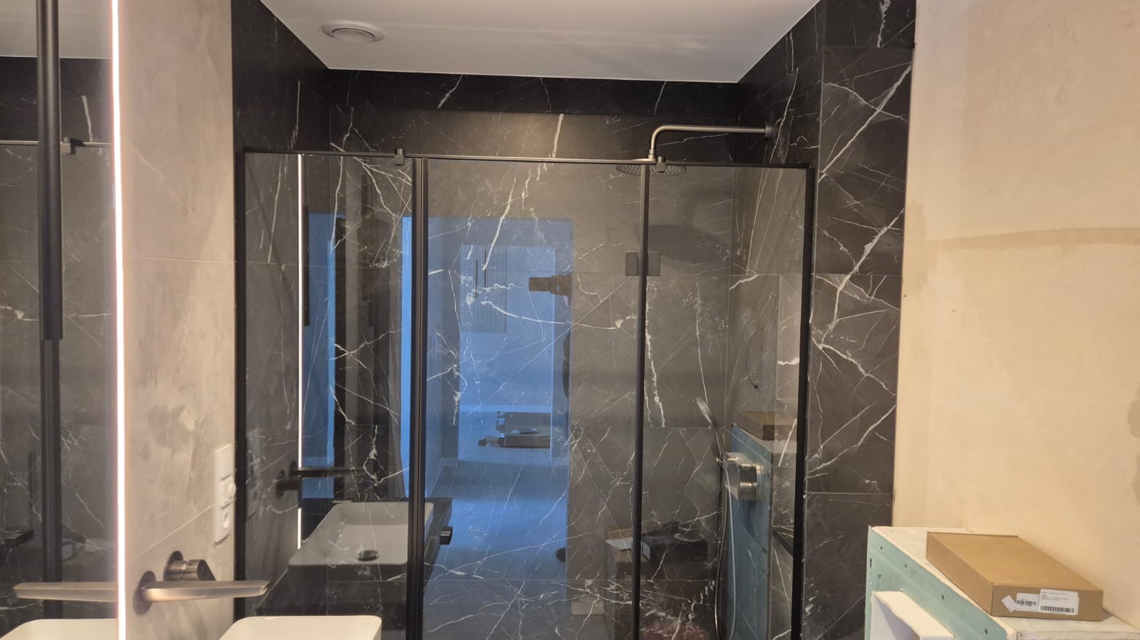 Nowoczesna łazienka z kabiną typu walk-in, wykończona płytkami imitującymi marmur. Widoczne oświetlenie LED i elementy armatury w odcieniach szarości. Częściowo wykończone ściany.