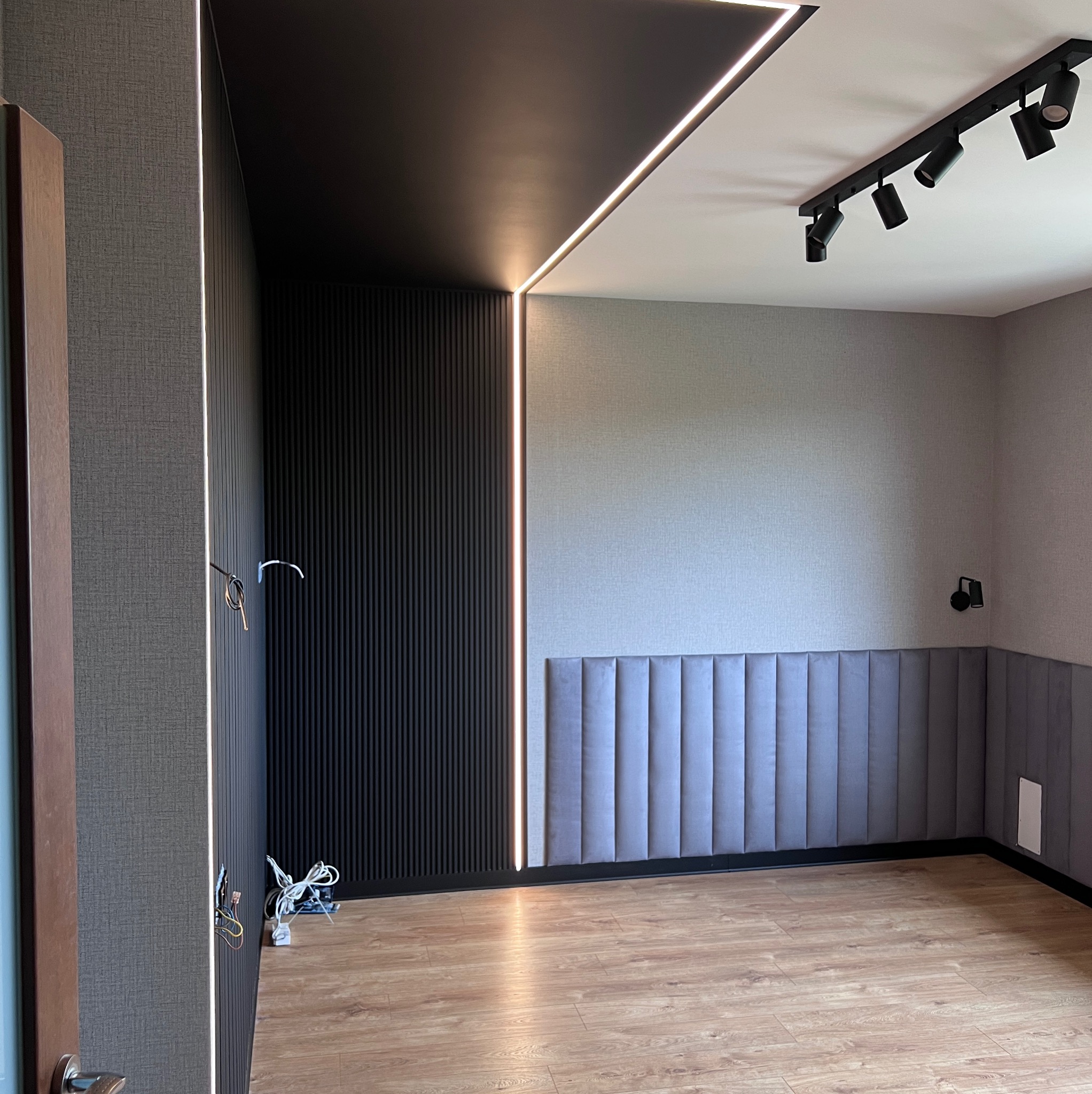 Nowoczesne wnętrze z czarną ścianą z lameli i podświetleniem LED, szara tapeta, panele podłogowe i oświetlenie punktowe na suficie. Częściowo widoczne kable.
