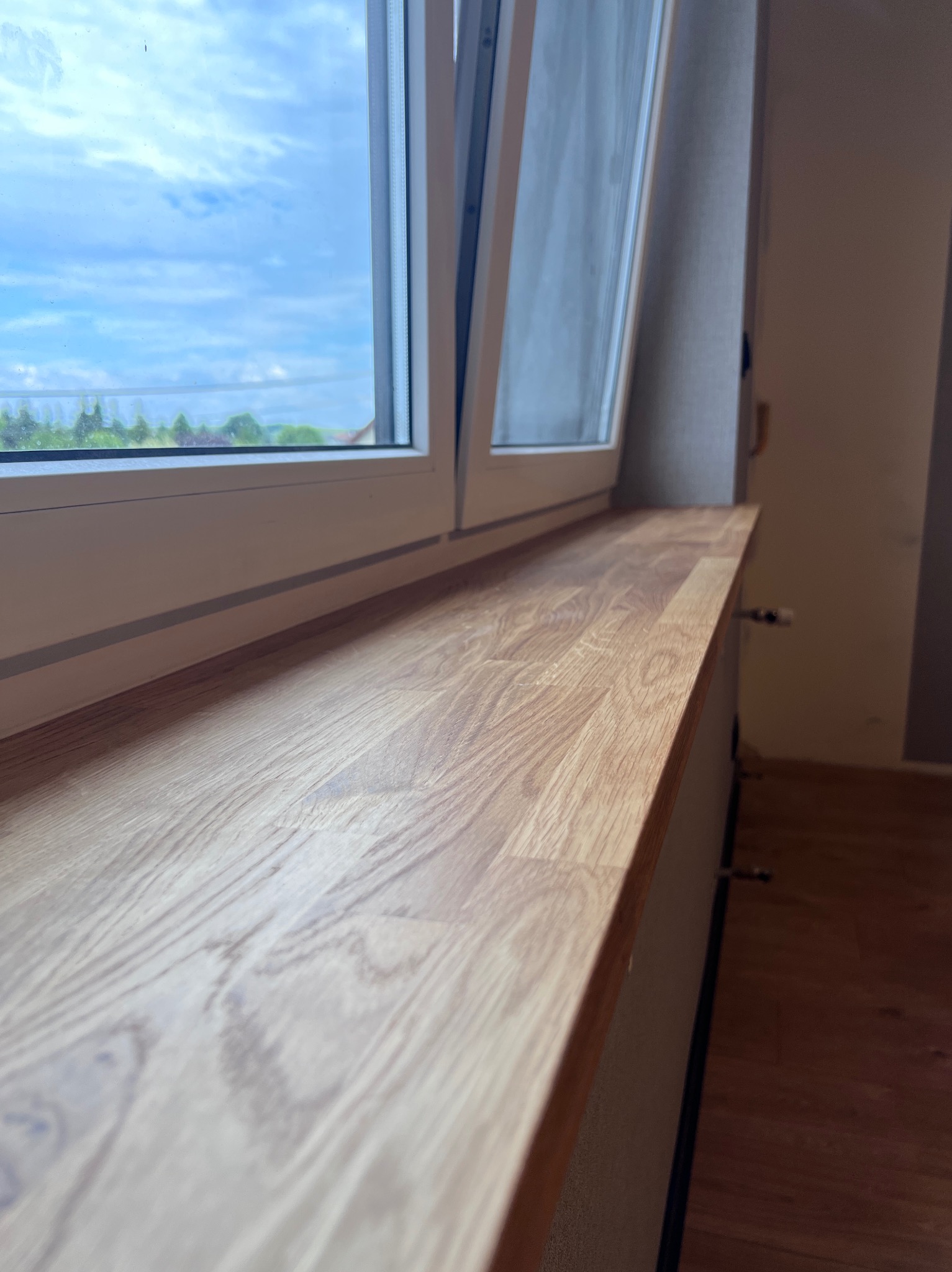 Zamontowany drewniany parapet pod oknem z widokiem na błękitne niebo i zieleń. Widoczny fragment grzejnika. Ujęcie z bliska, podkreślające strukturę drewna.