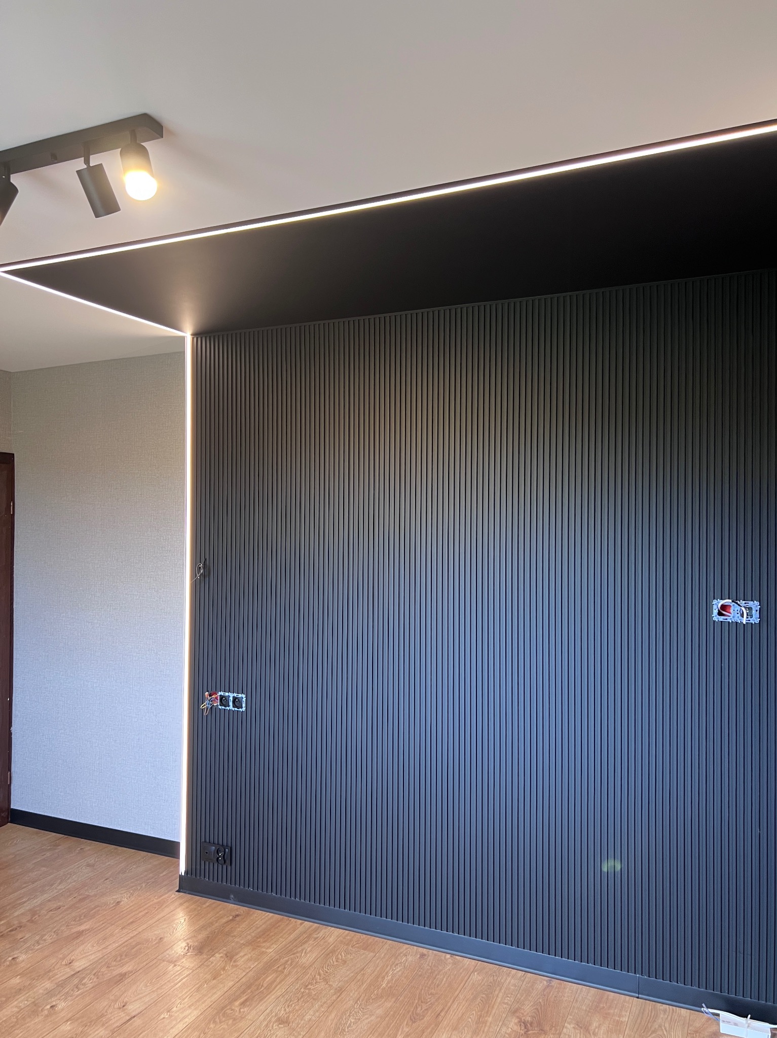 Nowoczesna ściana z lameli w kolorze antracytowym, oświetlenie LED na suficie i wzdłuż krawędzi, widoczne gniazdka elektryczne, podłoga z paneli.