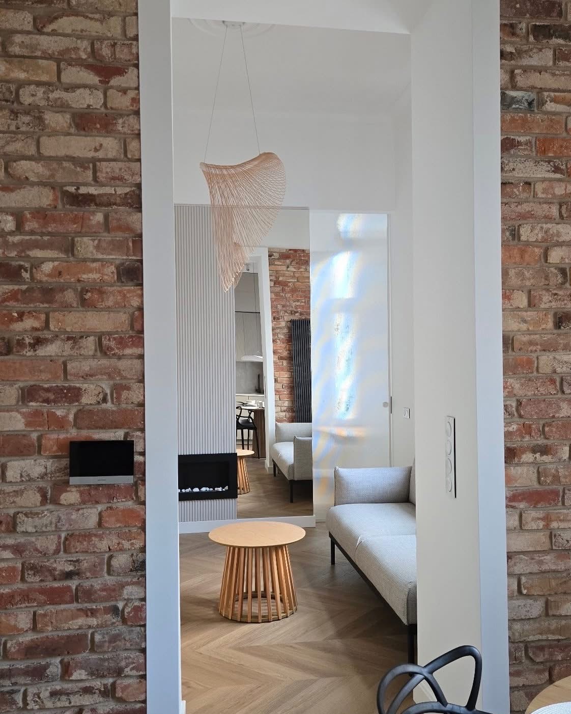 Salon po renowacji: ceglana ściana kontrastuje z nowoczesnym kominkiem i minimalistycznym oświetleniem. Jasna podłoga z parkietu w jodełkę.