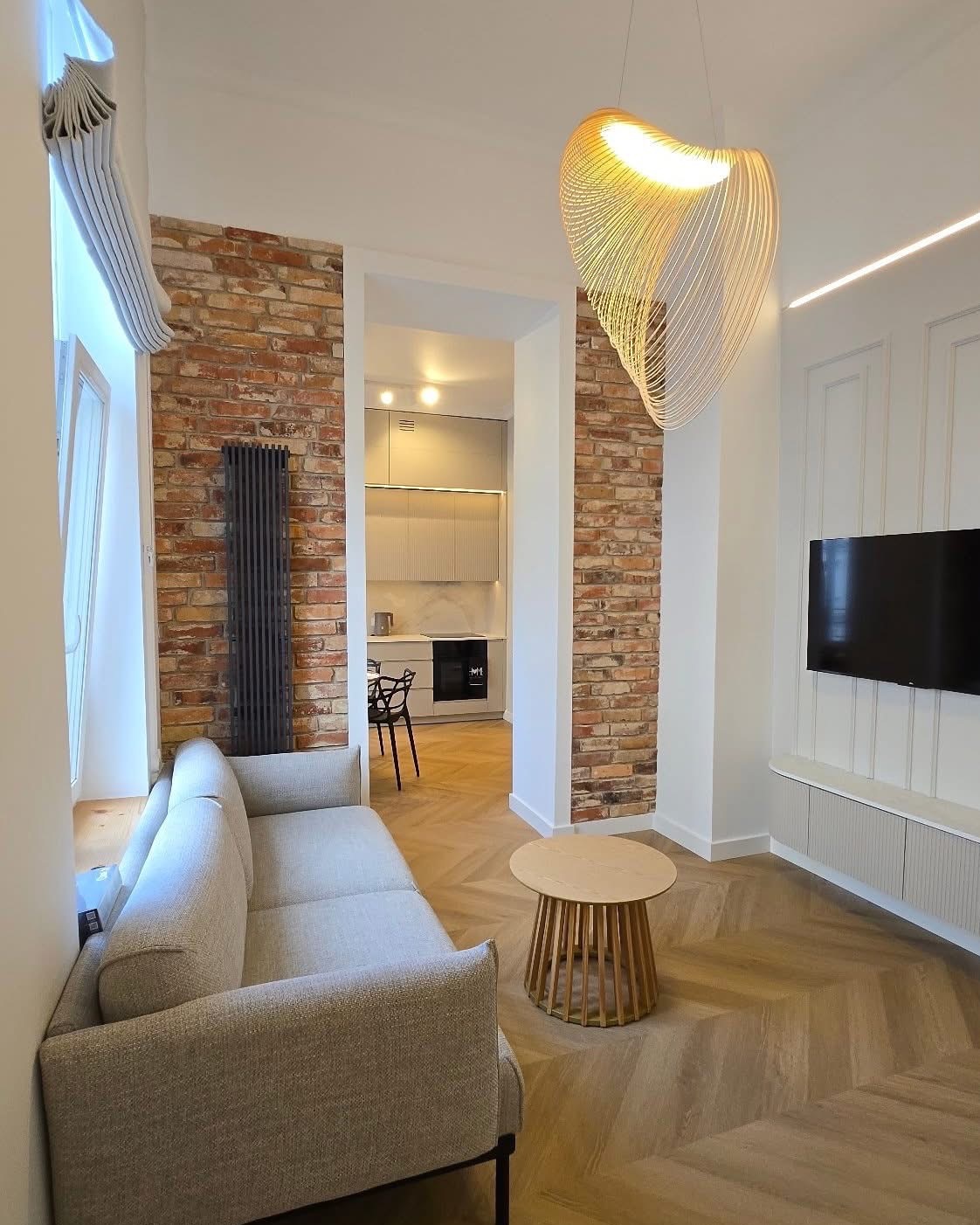 Stylowe wnętrze: szara sofa, parkiet w jodełkę, ceglana ściana z grzejnikiem i widok na kuchnię. Ozdobna lampa zwisa nad okrągłym stolikiem.