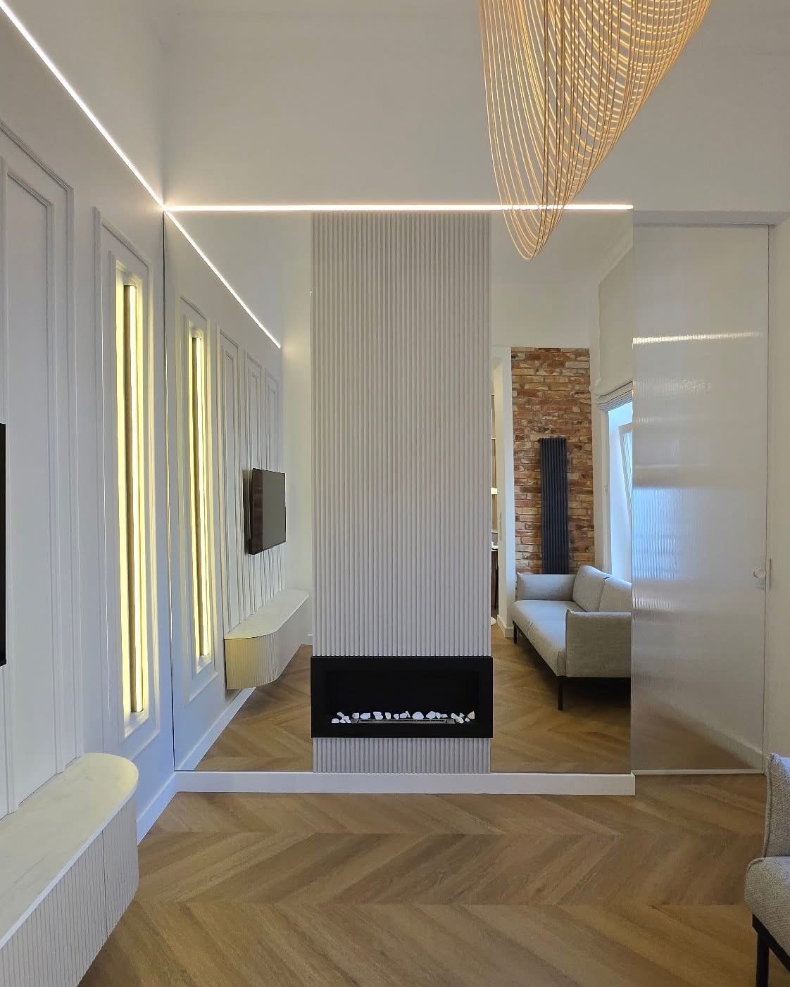 Nowoczesny salon z parkietem w jodełkę, ścianą z lamelami i minimalistycznym kominkiem. Oświetlenie LED podkreśla geometryczny design. Widok na drugą część pokoju.