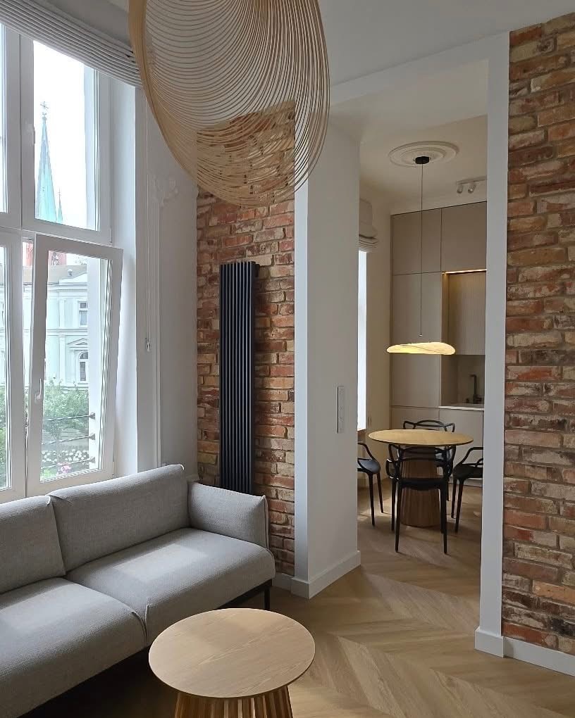 Stylowe wnętrze: salon z ceglaną ścianą, szarą sofą i drewnianym stolikiem kawowym. Widok na kuchnię z okrągłym stołem. Dekoracyjna lampa nad sofą.