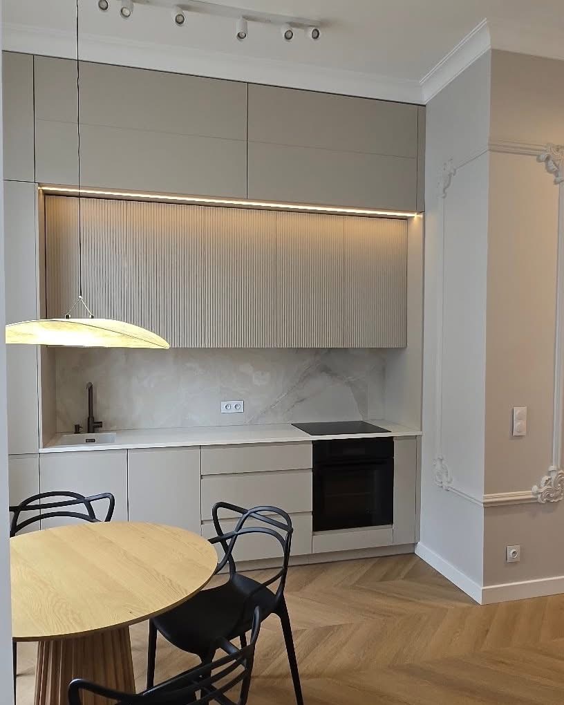 Nowoczesna kuchnia w beżach z drewnianym stołem i czarnymi krzesłami. Ryflowane fronty szafek górnych. Parkiet w jodełkę na podłodze. Dekoracyjne sztukaterie przy ścianie.