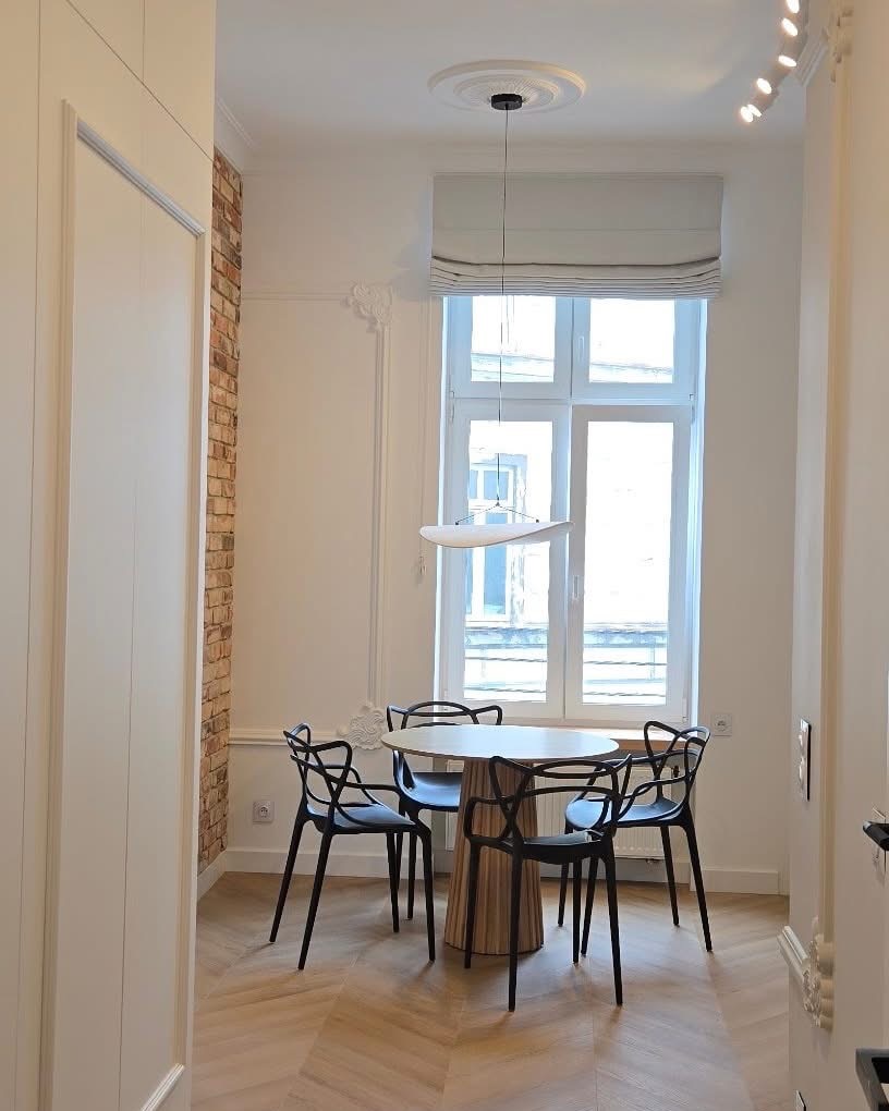 Jasne wnętrze po remoncie: okrągły stół z krzesłami, parkiet w jodełkę, ceglana ściana i okno z roletą rzymską. Nowoczesny design i wykończenie.