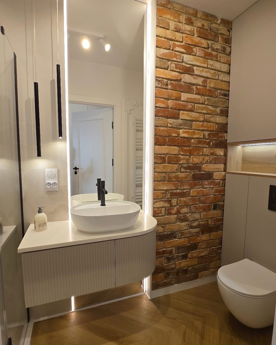 Nowoczesna łazienka z umywalką nablatową, lustrem z podświetleniem LED i ścianą z cegły. Parkiet w jodełkę, minimalistyczna armatura i ciepła kolorystyka.