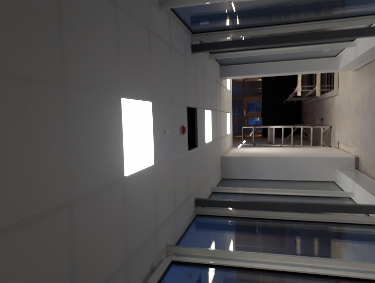 Wnętrze budynku z widokiem na sufit z oknami dachowymi, białe ściany z wbudowanym oświetleniem i drabina na górze.