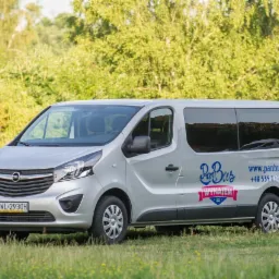 Srebrny Opel Vivaro z logo wypożyczalni w Warszawie, zaparkowany na trawiastym terenie z widocznym numerem rejestracyjnym i danymi kontaktowymi firmy.
