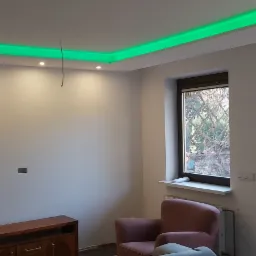 Nowoczesne wnętrze pokoju z podwieszanym sufitem z zielonym oświetleniem LED, szarymi ścianami, brązowymi oknami balkonowymi i drewnianą komodą.