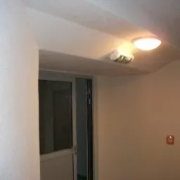 Wnętrze klatki schodowej z pomalowanymi ścianami na jasny kolor, widoczna lampa sufitowa, drzwi wejściowe i przełącznik.