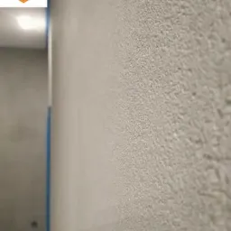 Ściana pokryta szarą, ziarnistą strukturą, zabezpieczona niebieską taśmą malarską przy suficie i podłodze, z widocznym logo Oferteo 'Najlepsi 2018' w rogu.