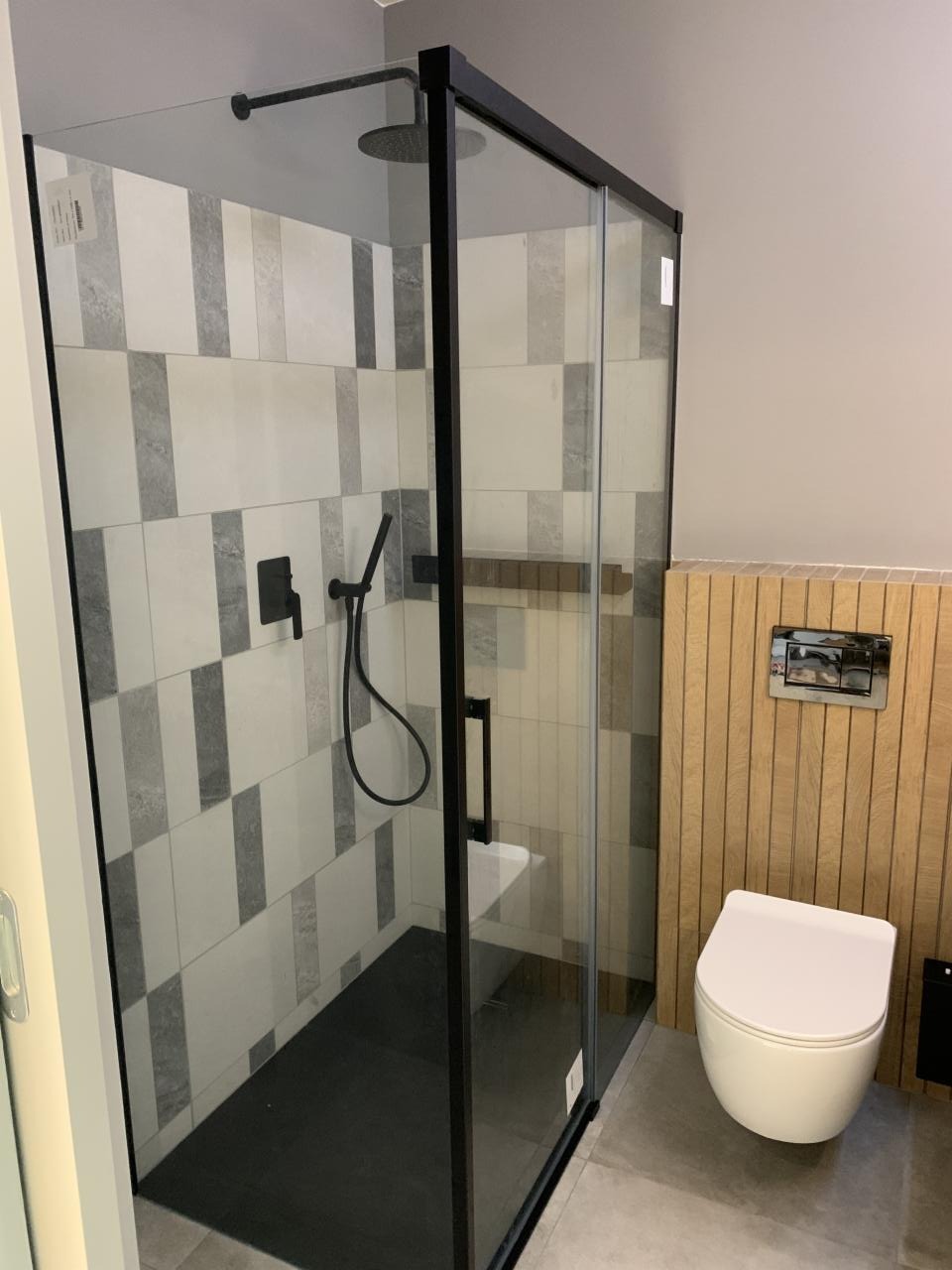 Nowoczesna kabina prysznicowa z czarną ramą i deszczownicą, wykończona płytkami w odcieniach szarości i bieli, obok toaleta wisząca i panel z drewnianą okładziną.
