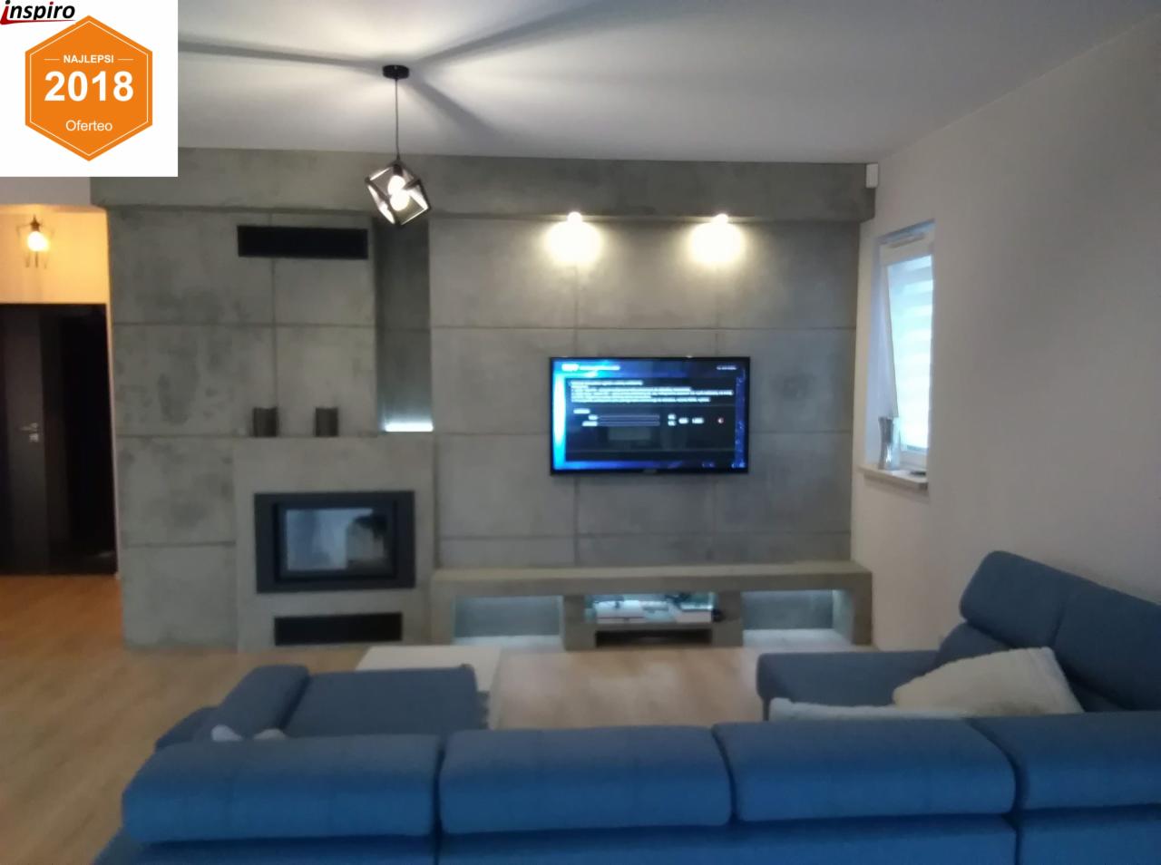 Salon z szarą ścianą z betonu architektonicznego, wnęką na kominek i telewizorem, oświetlony lampami punktowymi i wiszącą lampą designerską.