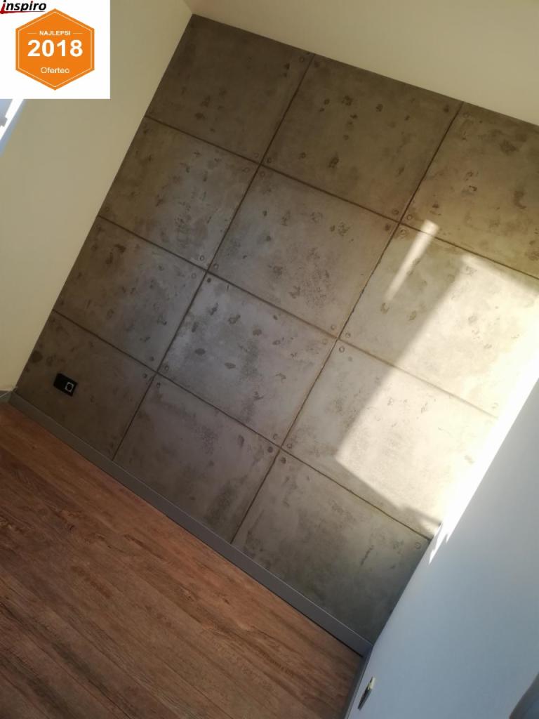 Ściana wykończona betonowymi płytami w kwadratowym wzorze, połączonymi metalowymi nitami, z widoczną podłogą z paneli drewnopodobnych i fragmentem białej ściany. Na górze widoczny fragment logo...