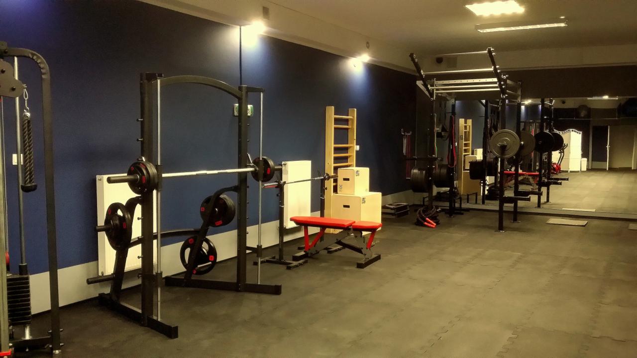 Profesjonalna siłownia z różnorodnym sprzętem do ćwiczeń siłowych, w tym suwnica Smitha z obciążeniami, drabinka gimnastyczna i skrzynie do plyometrii; widoczne odbicie w lustrze.