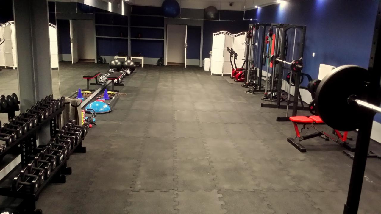 Przestronna siłownia z ciemną podłogą z puzzli, rzędem hantli po lewej stronie i różnorodnym sprzętem do ćwiczeń w tle, w tym maszyny siłowe, piłki do ćwiczeń i drabinki.