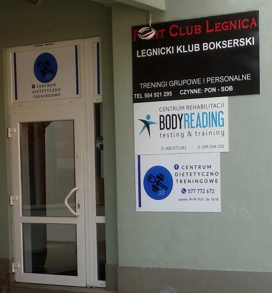 Wejście do centrum dietetyczno-treningowego z widocznymi tablicami informacyjnymi o ofercie, w tym treningach grupowych i personalnych oraz rehabilitacji.