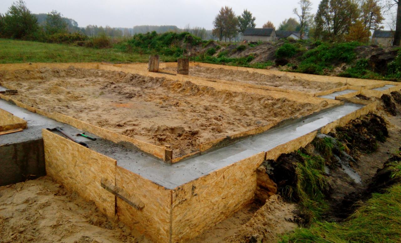 Wylane fundamenty pod budowę domu, widoczne drewniane szalunki i świeży beton, na tle zieleni i zabudowań.