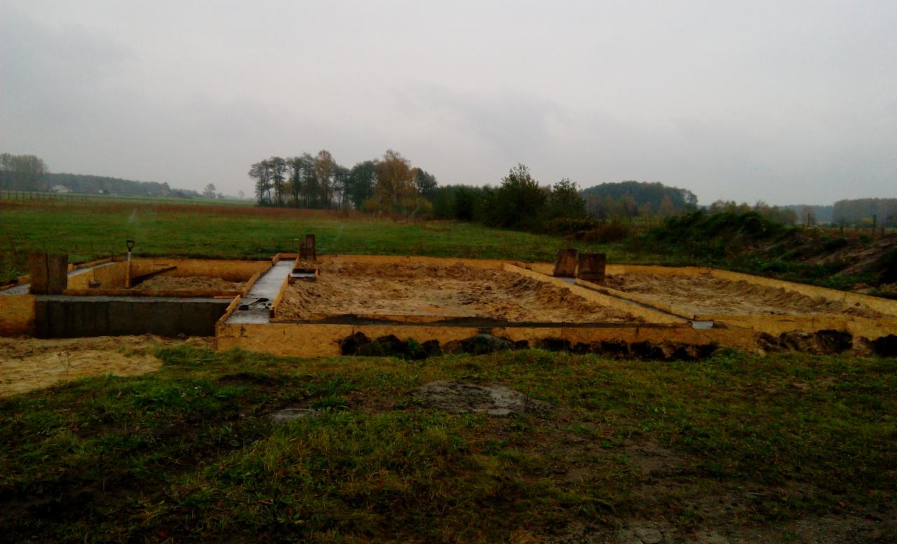 Fundamenty domu jednorodzinnego w trakcie budowy, widoczne szalunki i zbrojenie, w tle krajobraz wiejski w pochmurny dzień.