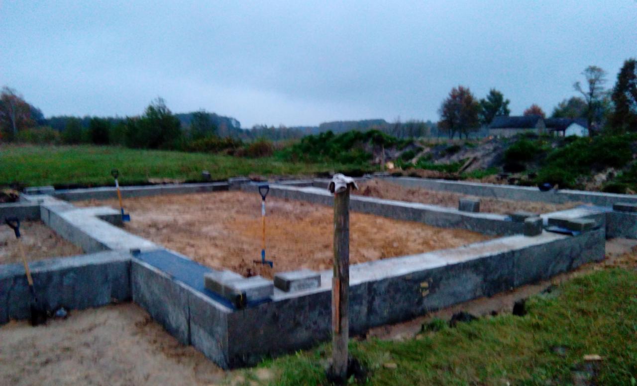 Fundamenty domu jednorodzinnego z widocznymi łopatami i drewnianym słupkiem w tle krajobraz z drzewami na pochmurnym niebie.