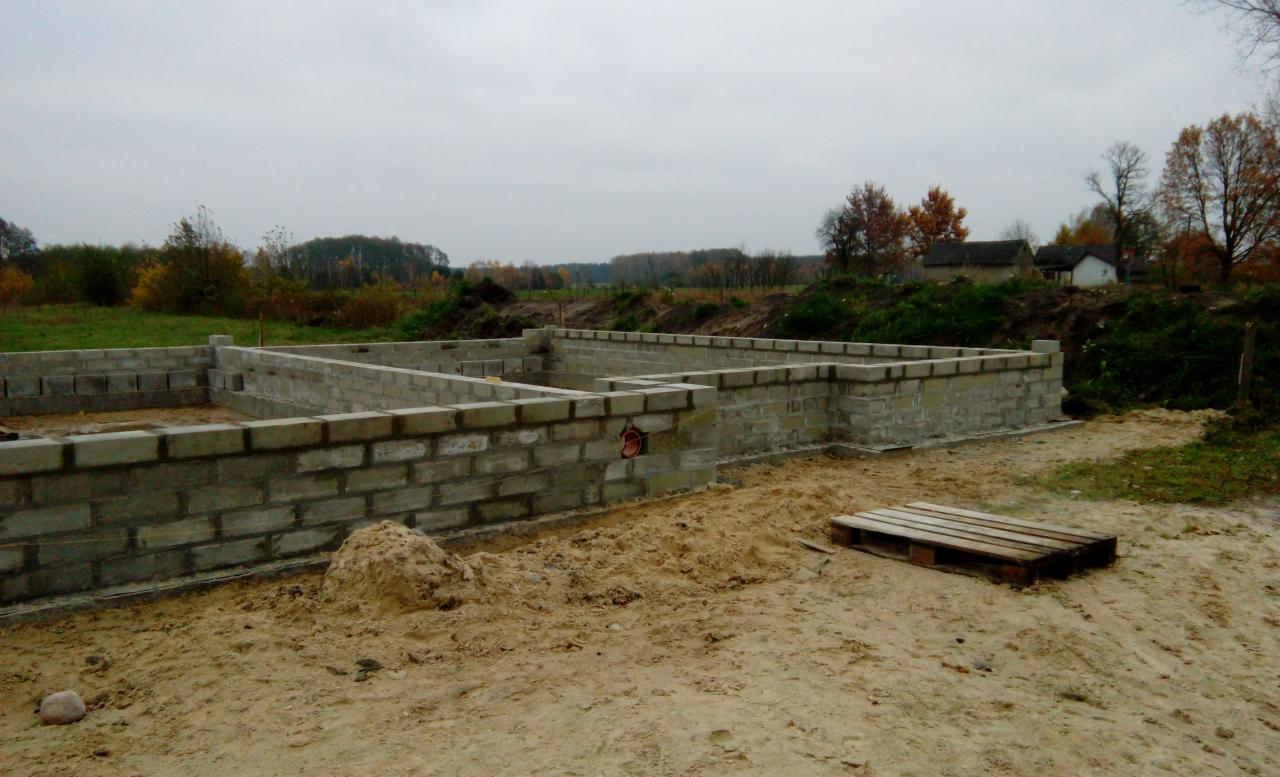 Fundamenty domu z bloczków betonowych, widoczna skrzynka elektryczna w ścianie, teren piaszczysty, w tle drzewa i zabudowania.