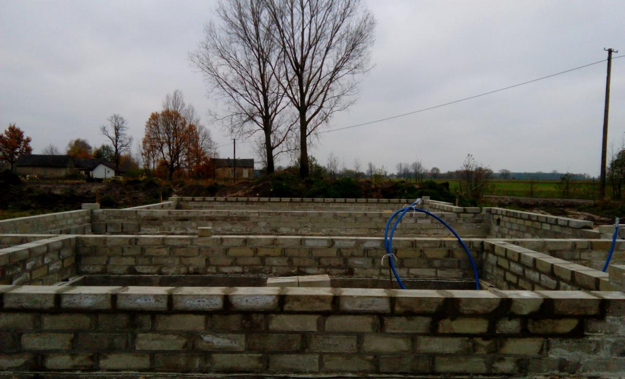 Fundamenty domu z bloczków betonowych, widoczne niebieskie rury instalacyjne, w tle drzewa i zabudowania wiejskie, pochmurne niebo.