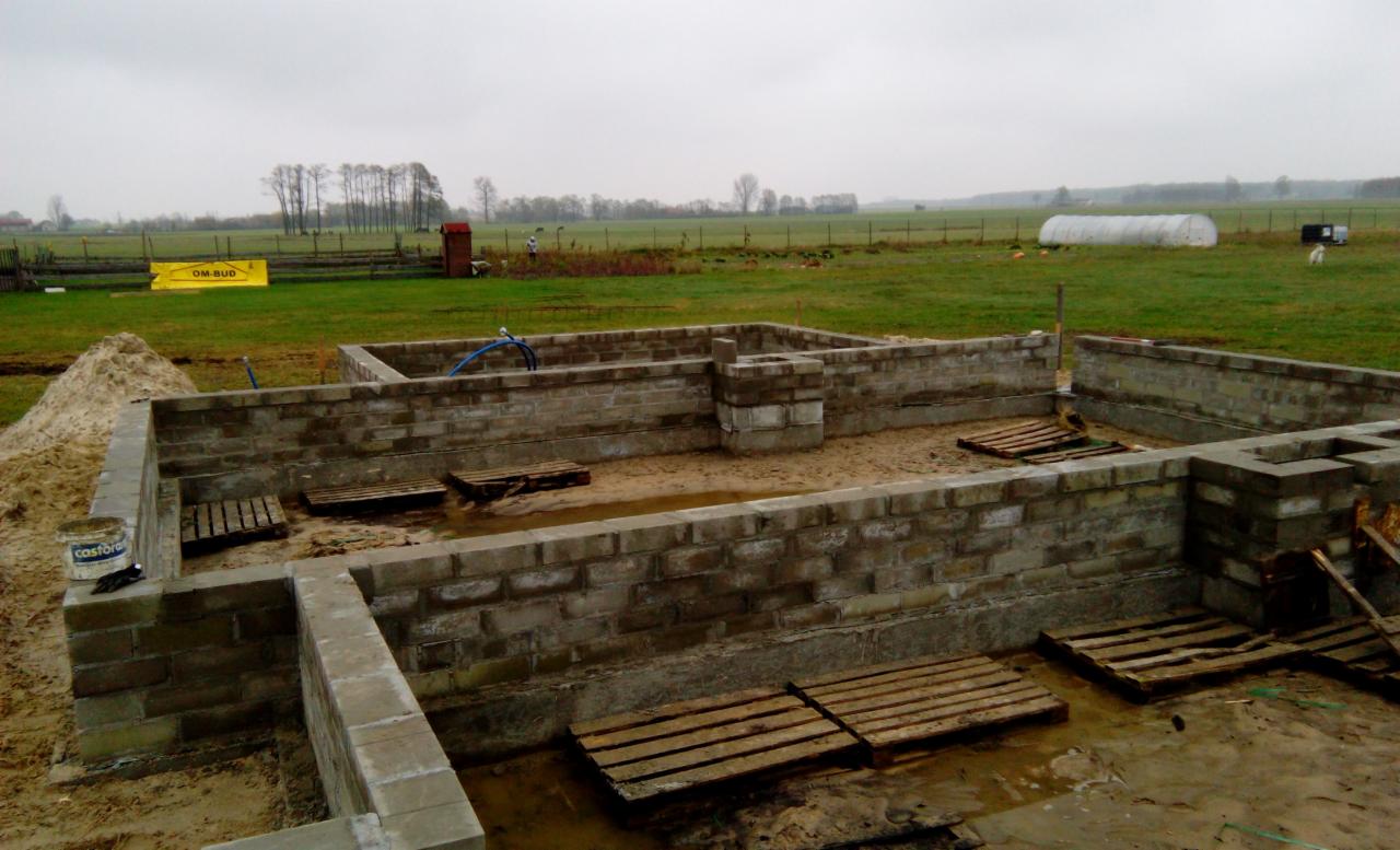 Fundamenty domu z bloczków betonowych na piaszczystym gruncie, widoczne drewniane palety wewnątrz fundamentów, w tle zielone pole i pochmurne niebo.